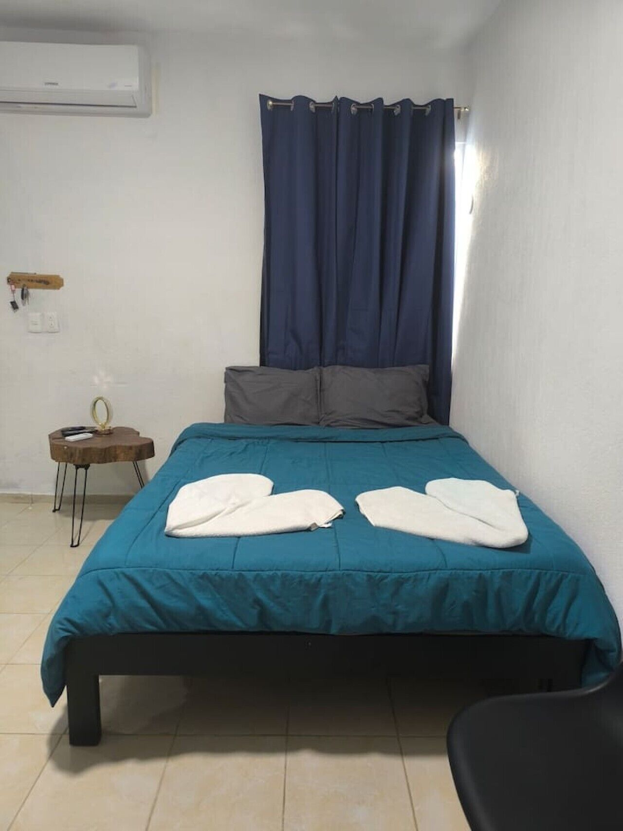 A single bed with a teal bedspread, two white pillows, a dark blue curtain, a small wooden side table with a lamp, and a black chair.