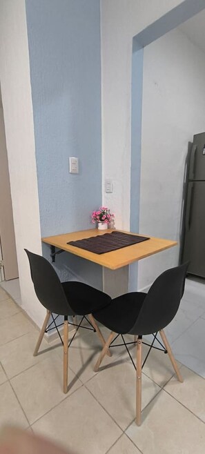 A small kitchen with a wooden table and two black chairs, a refrigerator, and a wall with a light switch. - Loft acogedor y funcional Facturamos (Merida)