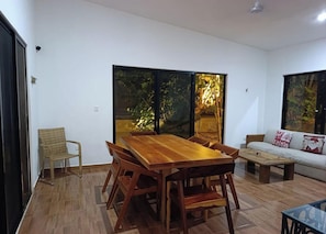A dining area with a wooden table and chairs, a sofa, and a window with a view of greenery.