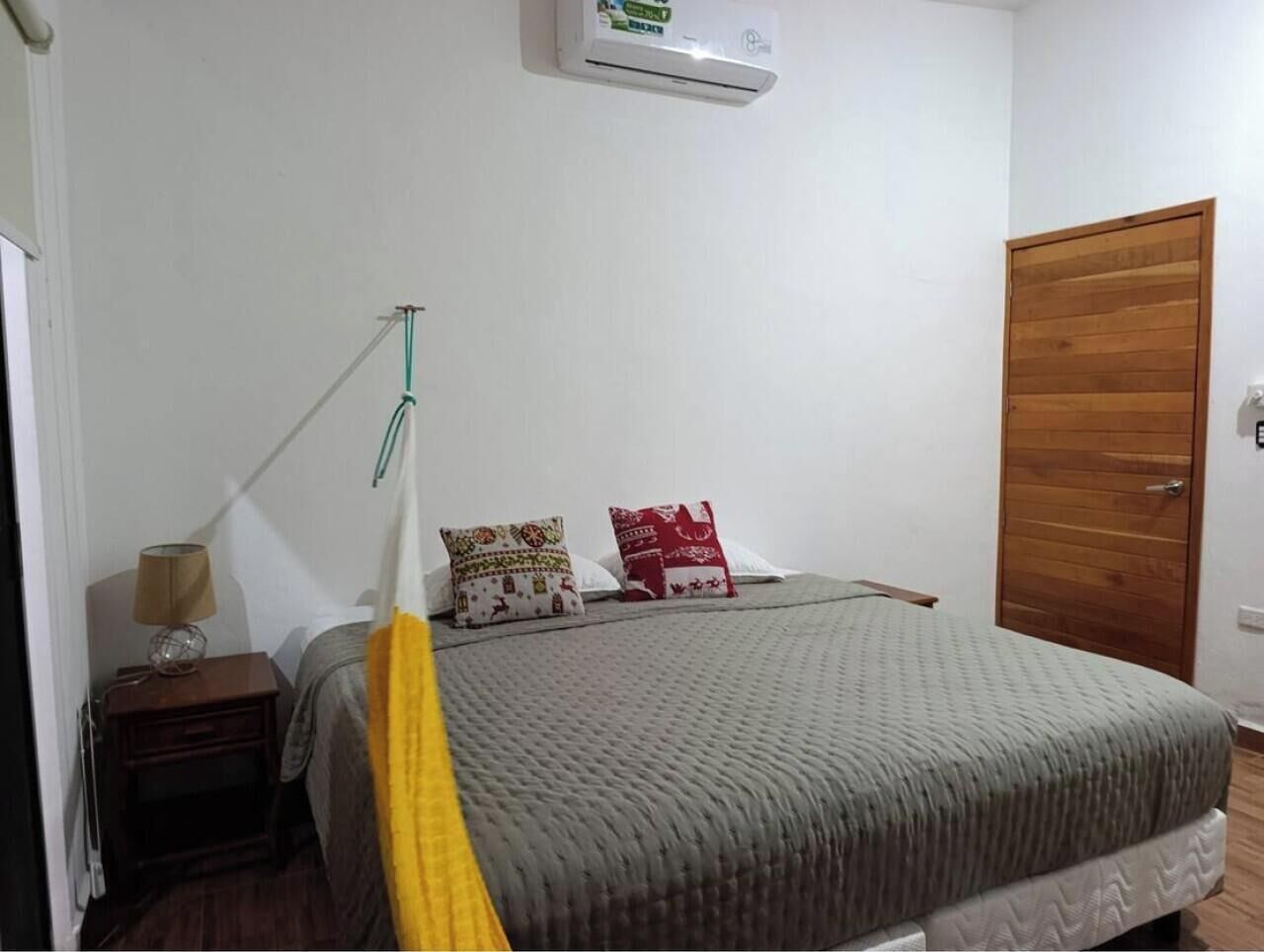 A bedroom with a bed, a nightstand, a lamp, a wall-mounted air conditioner, and a wooden door.
