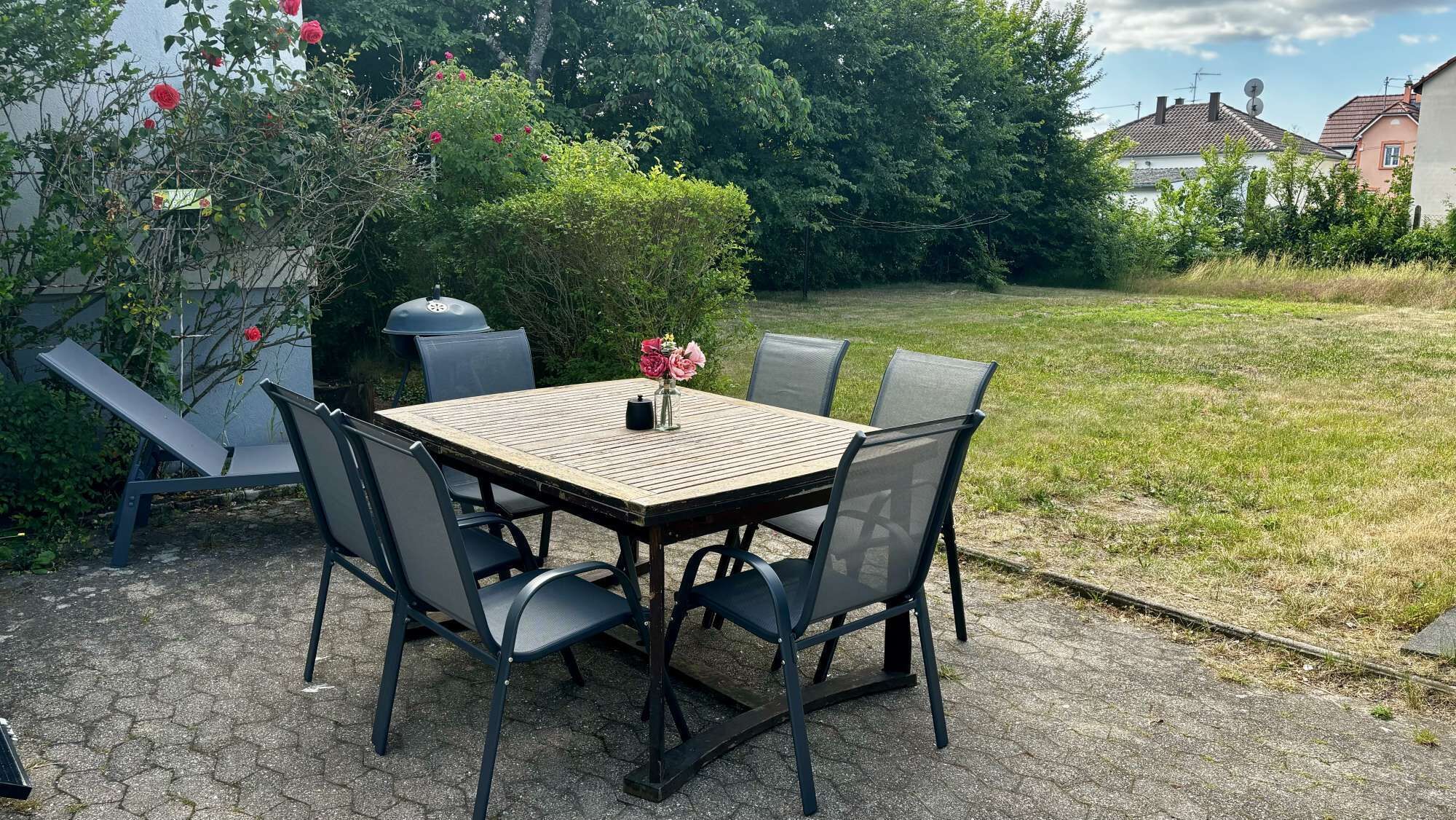 Un patio exterior con una mesa cuadrada, cuatro sillas, una parrilla y un jarrón con flores.