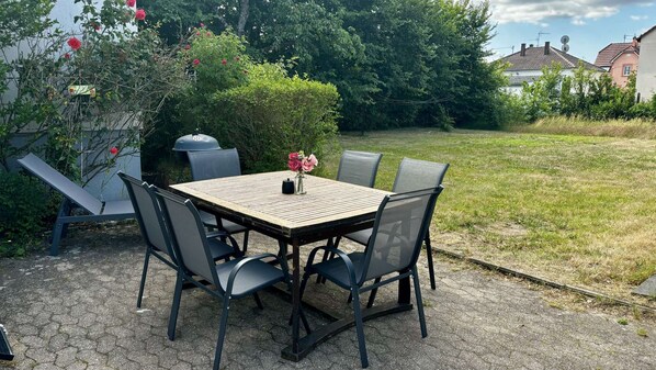Une terrasse extérieure avec une table carrée, quatre chaises, un barbecue et un vase rempli de fleurs.