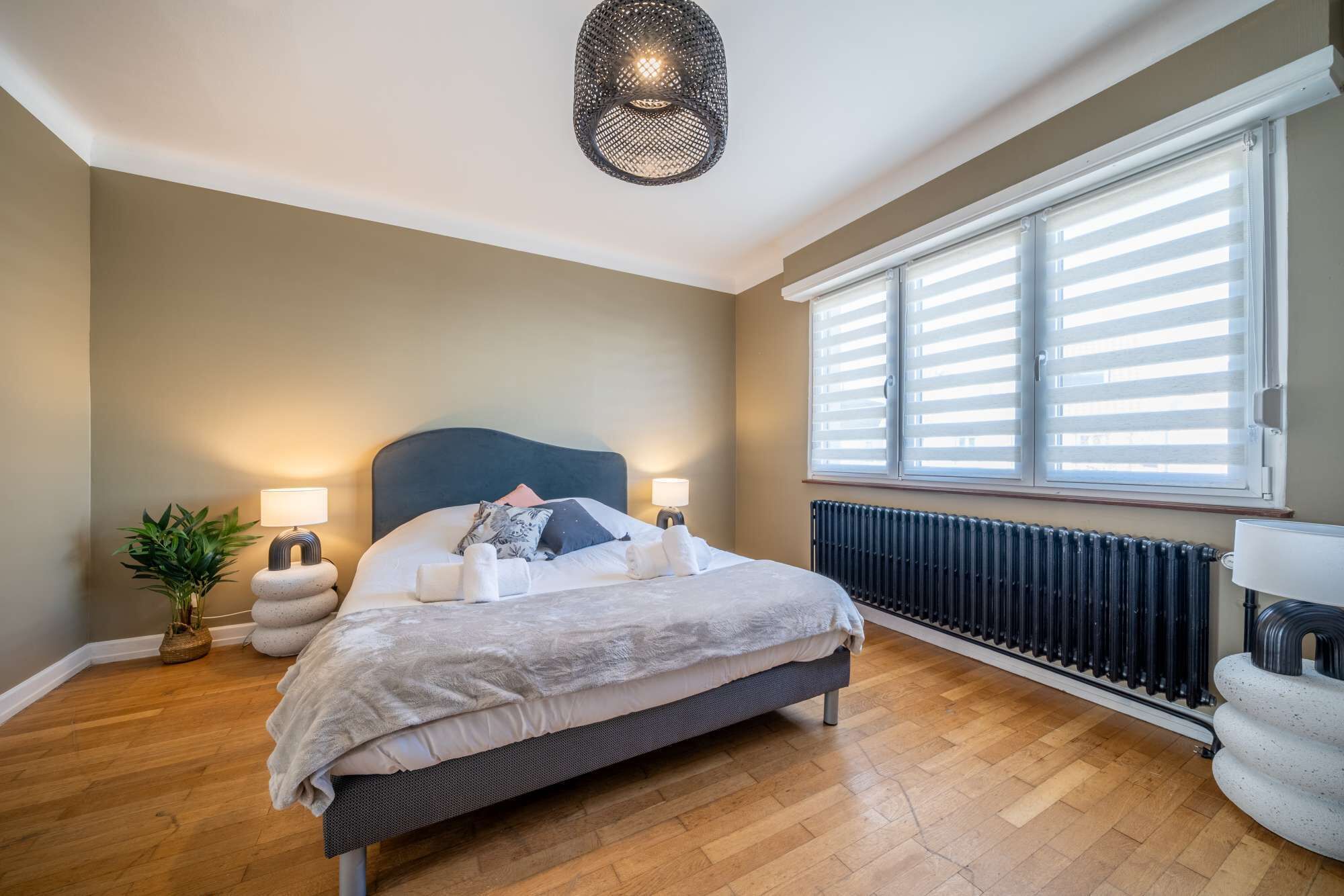 A bedroom with a bed, bedside lamps, a radiator, and a window with blinds.