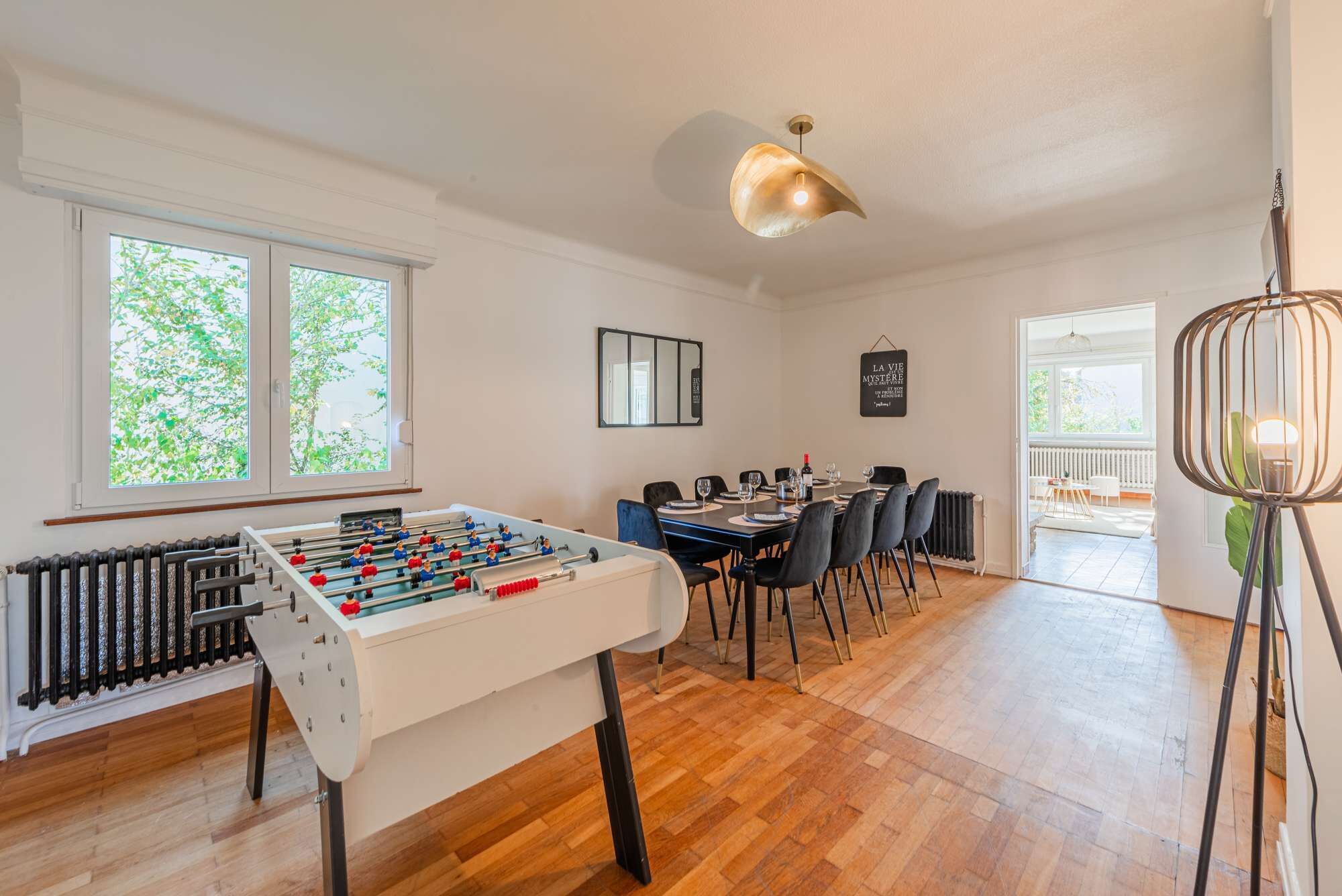Ett rum med ett fotbollsspel, ett matbord med stolar, ett element och ett stort fönster med utsikt över träd.