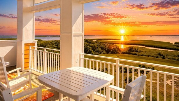A balcony with a table and chairs, overlooking a sunset view of the ocean and landscape.