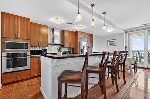 A modern kitchen with wooden cabinets, stainless steel appliances, a bar area with stools, and a view of the outdoors. - New! Oceanfront Penthouse/ 2 Decks/ Pool/Beach/Gym (Virginia Beach)