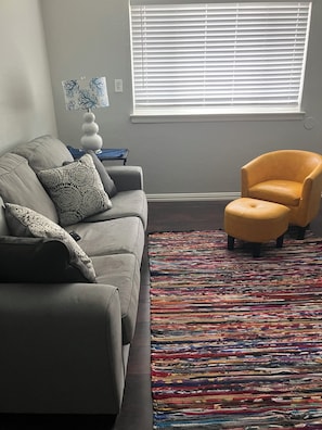 Ein Wohnzimmer mit grauem Sofa, einem gelben Sessel, einem bunten Teppich und einem Fenster mit Jalousien.