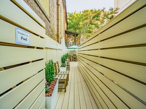 Eine schmale Holzterrasse mit Bänken und Pflanzkübeln, ein Schild, das einen Pool anzeigt, und ein Backsteinhaus im Hintergrund.