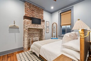 Une chambre avec une cheminée en briques, un lit avec une literie blanche, un plancher en bois, un tapis à carreaux, une table de chevet, une lampe et une fenêtre avec un store.
