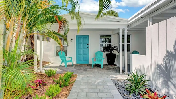 Ein weißes Gebäude mit einer blauen Tür, zwei türkisblauen Stühlen und verschiedenen Pflanzen.