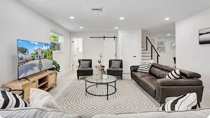 Ein modernes Wohnzimmer mit einem Flachbildfernseher, einer Couch, zwei Sesseln, einem Couchtisch und einer Treppe im Hintergrund.