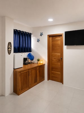 A room with a wooden cabinet, a coffee maker, a blue lamp, a potted plant, a wall-mounted TV, and a door with a wooden frame.