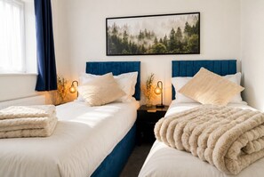 Habitación de hotel con dos camas, una mesita de noche con lámpara, un cuadro con un paisaje forestal y una ventana con cortinas.