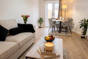 Ein modernes Wohnzimmer mit einer Couch, einem Couchtisch mit Kerze, einer Obstschale und einem Essbereich mit Stühlen und einem Tisch.
