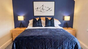 Una cama con un edredón azul oscuro, dos lámparas de noche, una mesita de noche de madera, un cuadro enmarcado en la pared y un suelo alfombrado.