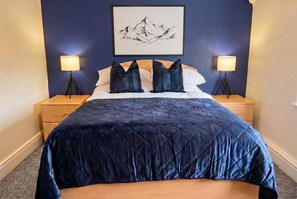 A bed with a dark blue quilt, two bedside lamps, a wooden nightstand, a framed picture on the wall, and a carpeted floor. - Pool Table & Free Parking · 4-Bed Home Sleeps 8 · Cowley, Oxford (Oxfordshire)