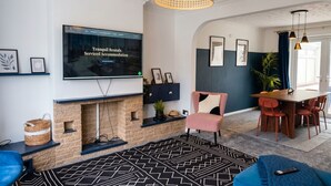 A living room with a fireplace, a television mounted on the wall, a dining area with a table and chairs, and a blue sofa. - 4-Bed Headington Home · Sleeps 8 with Parking · Walk to JR Hospital (Headington)