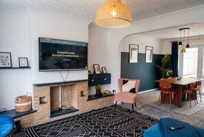 Ein Wohnzimmer mit Kamin, einem an der Wand montierten Fernseher, einer Essecke mit Tisch und Stühlen sowie einem blauen Sofa.