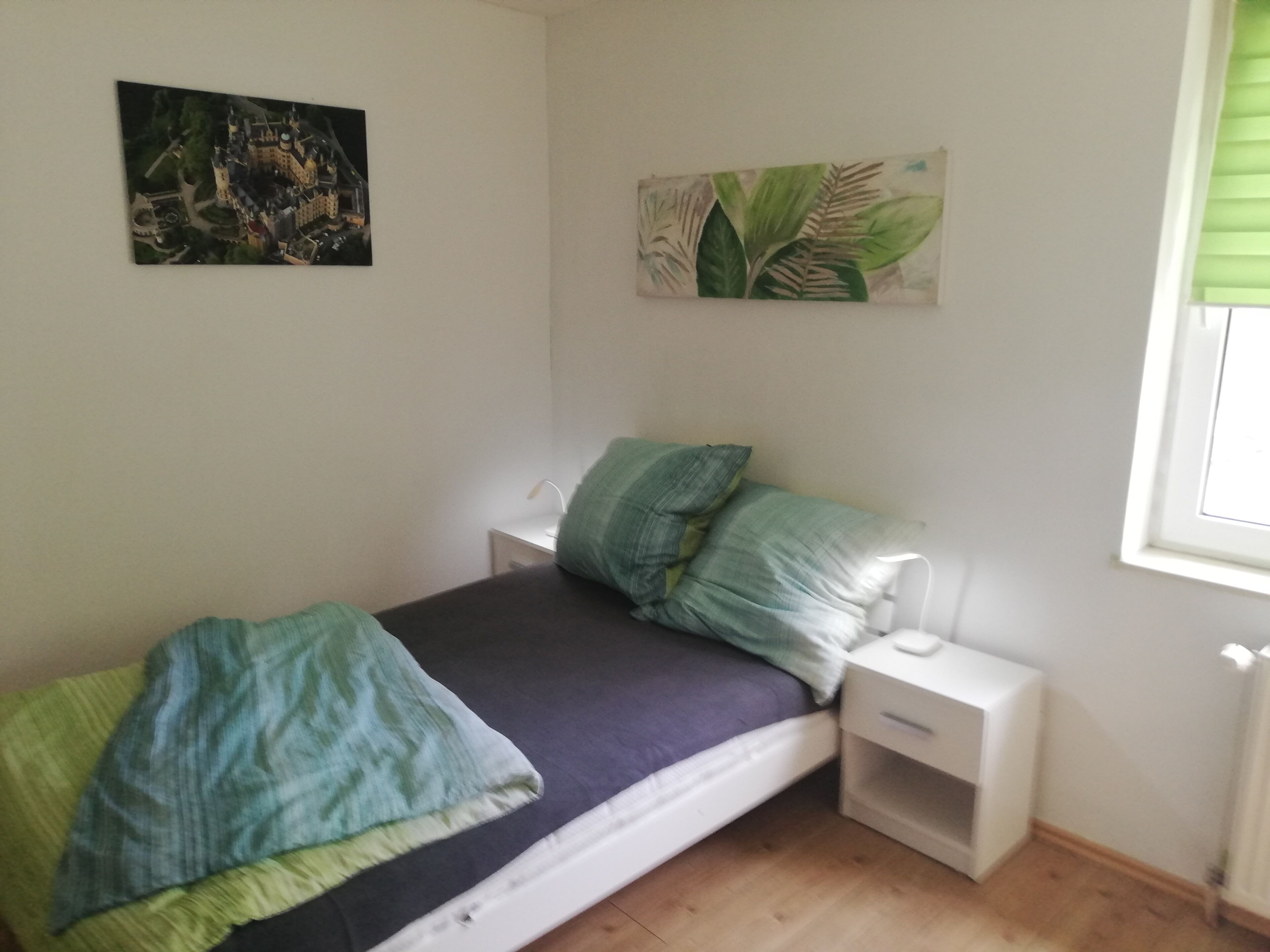 Une chambre à coucher comprenant un lit, une table de chevet, une lampe, une fenêtre avec des stores et deux œuvres d’art murales.