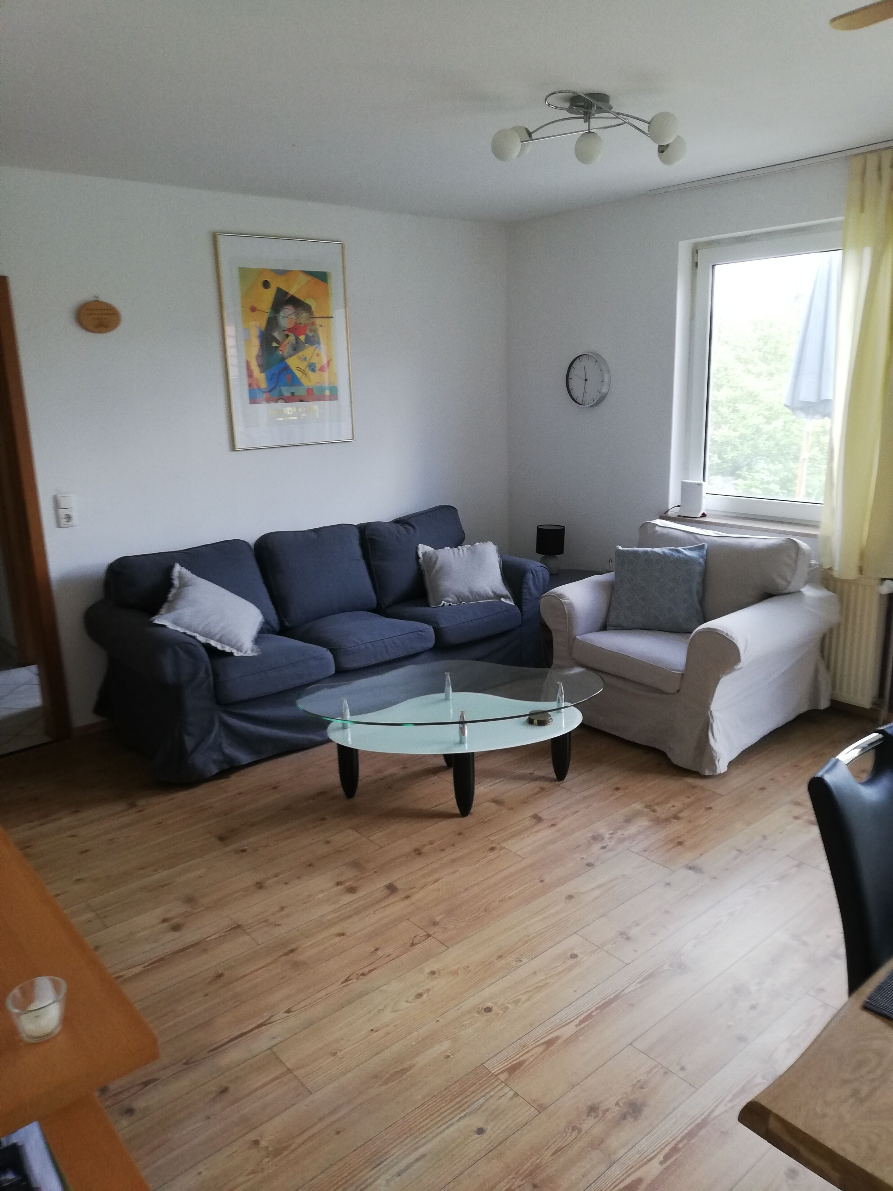 A living room with a blue sofa, a beige armchair, a glass coffee table, wooden flooring, a ceiling fan, a clock, and a window with curtains.