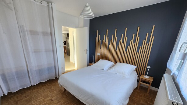 A bedroom with a bed, wooden floor, white curtains, a wall-mounted light fixture, and a radiator. - Holiday Home 'Gîte Rural Le Vendangeoir' with Private Garden and Wi-Fi (Colombé-la-Fosse)