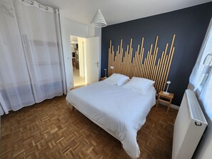 Et soveværelse med en seng, trægulv, hvide gardiner, en lampe på væggen og en radiator.