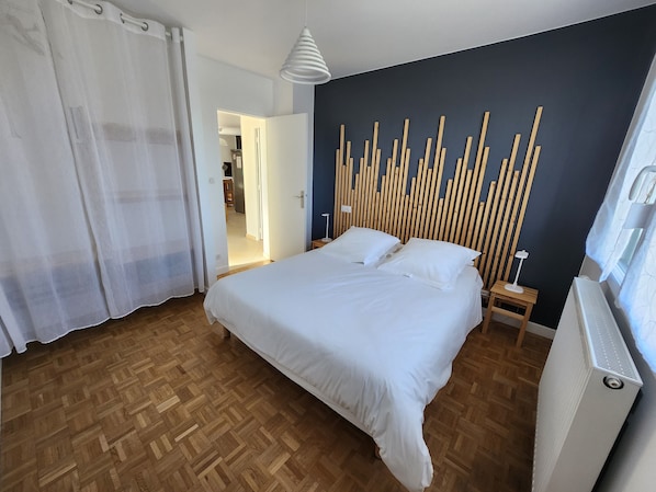 A bedroom with a bed, wooden floor, white curtains, a wall-mounted light fixture, and a radiator.