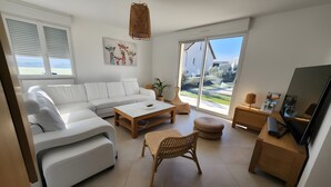A modern living room with a white sofa, a coffee table, a wicker chair, a TV, and a large window with a view. - Holiday Home 'Gîte Rural Le Vendangeoir' with Private Garden and Wi-Fi (Colombé-la-Fosse)