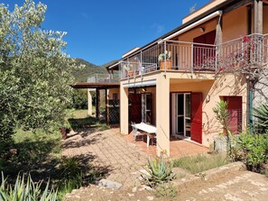 Una casa de dos pisos con un balcón, un patio con mesa y sillas, y un jardín con diversas plantas.