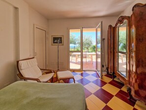 Quarto com piso xadrez, uma cama, duas cadeiras, uma mesa e vista para área externa através de porta de vidro, onde se vê mesa e cadeiras.