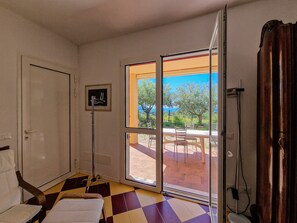 Quarto com chão quadriculado, porta, quadro na parede e vista para área externa com mesa e cadeiras através de uma porta de vidro.