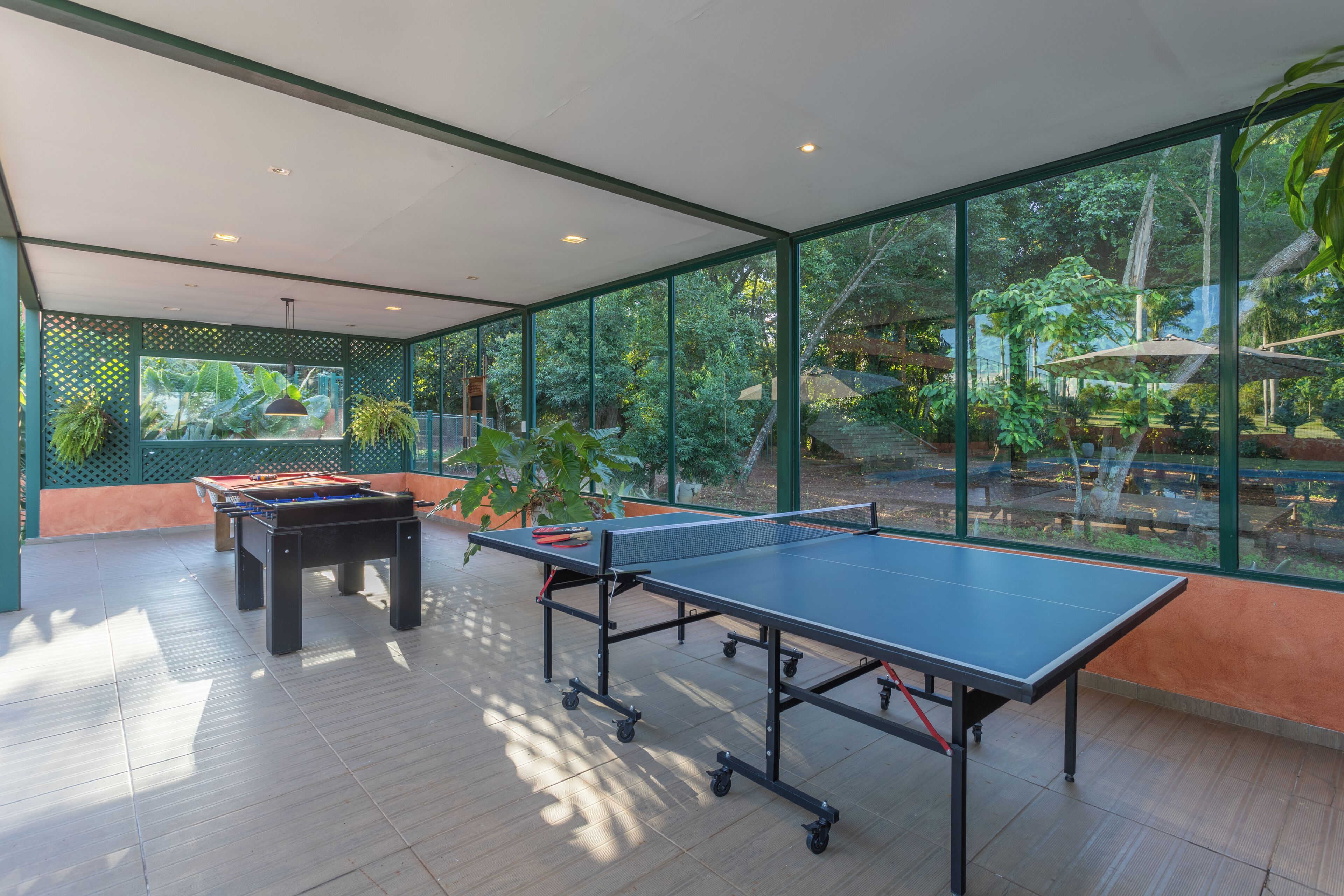 An indoor ping pong table and foosball table in a room with large windows and a view of a garden.