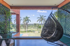 A balcony with a hanging chair, a view of a grassy area with palm trees, and a lattice wall with plants.