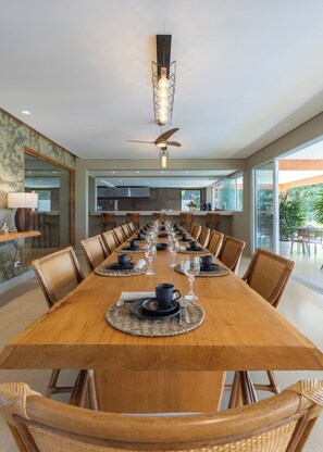 Un comedor con una larga mesa de madera dispuesta para una comida, rodeada de sillas de mimbre, con un ventilador de techo y luces colgantes.