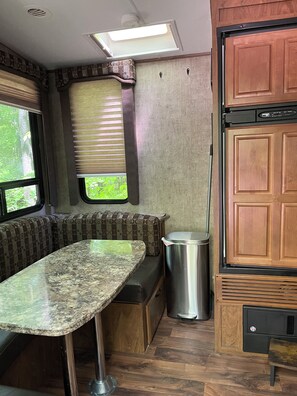 A compact RV kitchen with a built-in stove, a sink, a window with blinds, and a bench seating area.