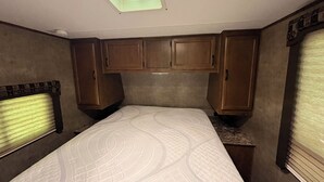 Ein Schlafzimmer mit Oberlicht, einem Bett mit gemustertem Überwurf, Holzschränken und einem Fenster mit Jalousien.