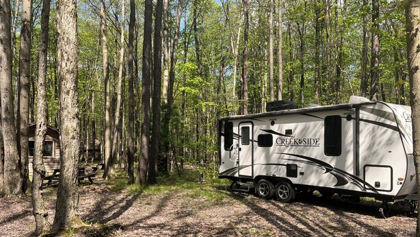 Ein weißer Wohnmobil mit 'CREEKSIDE' Schriftzug, geparkt in einer bewaldeten Gegend mit einem Picknicktisch und einem Haus im Hintergrund.
