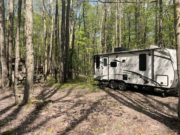 A white RV with 'CREEKSIDE' branding parked in a forested area with a picnic table and a cabin in the background.