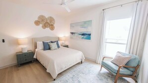 Una camera da letto con un letto, comodini, una sedia, una ventola a soffitto, una finestra con tende e un quadro appeso al muro.