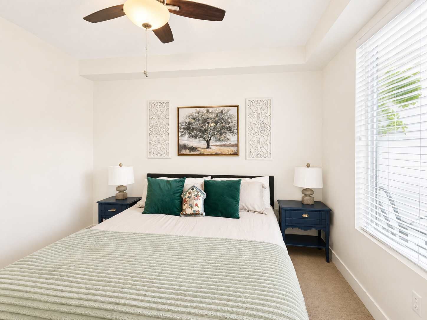 A bedroom with a bed, two nightstands, a ceiling fan, a window with blinds, and two wall-mounted decorative panels.