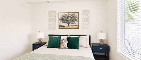 A bedroom with a bed, two nightstands, a ceiling fan, a window with blinds, and two wall-mounted decorative panels.