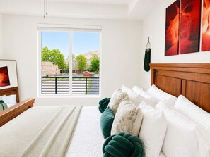 Une chambre avec une grande fenêtre, un lit agrémenté d’oreillers blancs et verts, une tête de lit en bois et un tableau accroché au mur.