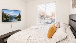 Un dormitorio con cama, televisor, ventana y vistas a un lago y montañas.