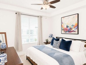 Ein Schlafzimmer mit Bett, Deckenventilator, Fenster mit Vorhängen, Nachttisch mit Lampe und einem gerahmten abstrakten Gemälde.