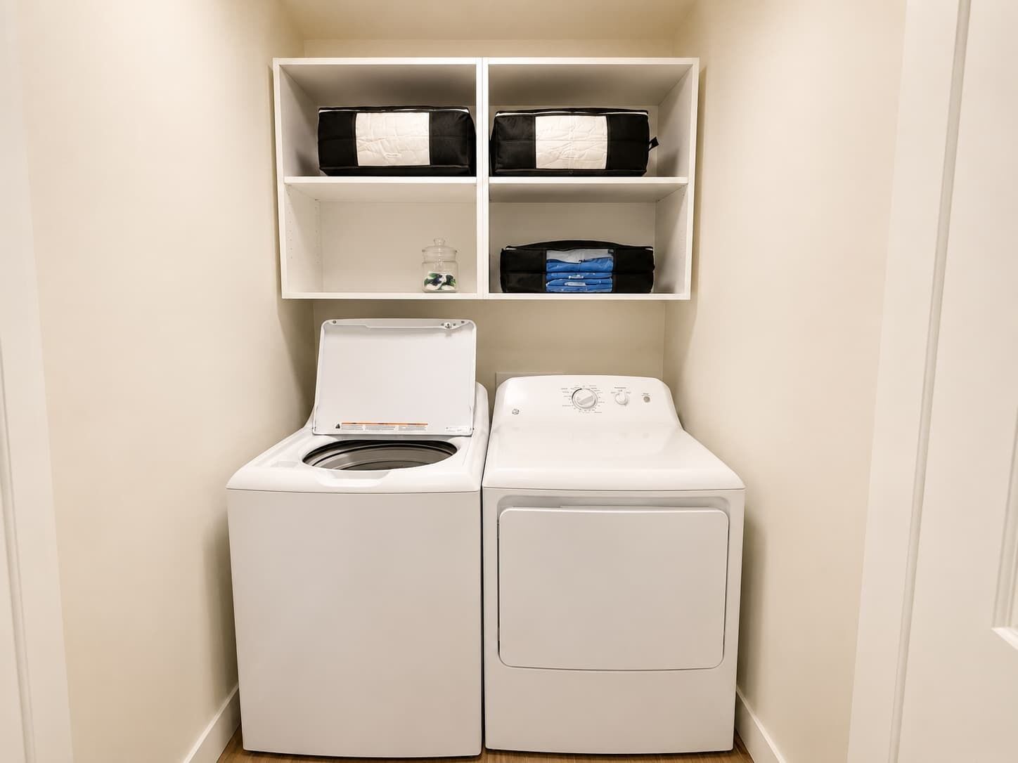 A laundry room with two white washing machines, a shelf above with storage boxes, and a glass jar.