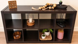 A dark wooden console table with multiple shelves, a small wooden box, a candle, a plant, and a remote control. - 2BR Urban Oasis in Downtown SLC - Sleeps 6, Gym (Salt Lake City)