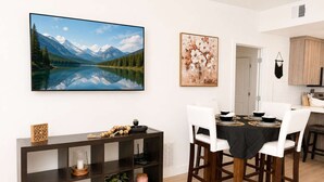 A dining area with a table set for four, a large landscape painting, a console table with decorative items, and a kitchen area in the background. - 2BR Urban Oasis in Downtown SLC - Sleeps 6, Gym (Salt Lake City)