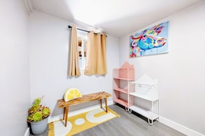 A room with a bench, a potted plant, a colorful painting, a metal shelf unit, and a window with beige curtains. - Fresh 3BR East Bay Home near OAK 2 Parking Spots (San Leandro)