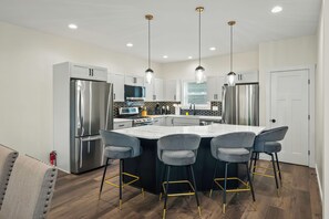 Een moderne keuken met roestvrijstalen apparatuur, een centraal eiland met barstoelen, hanglampen en uitzicht op de buitenlucht door een raam.