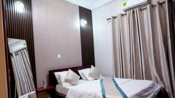 A hotel room with a bed, a bedside table, a mirror, and a window with curtains. - Clos D'or appartement (Douala)